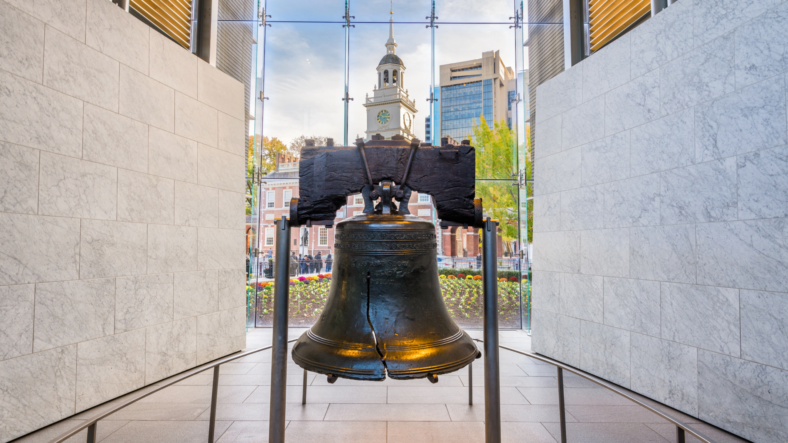 The Liberty Bell displayed inside a glass-walled pavilion, with its iconic crack visible, framed by marble walls and windows looking out toward historic buildings in Philadelphia.